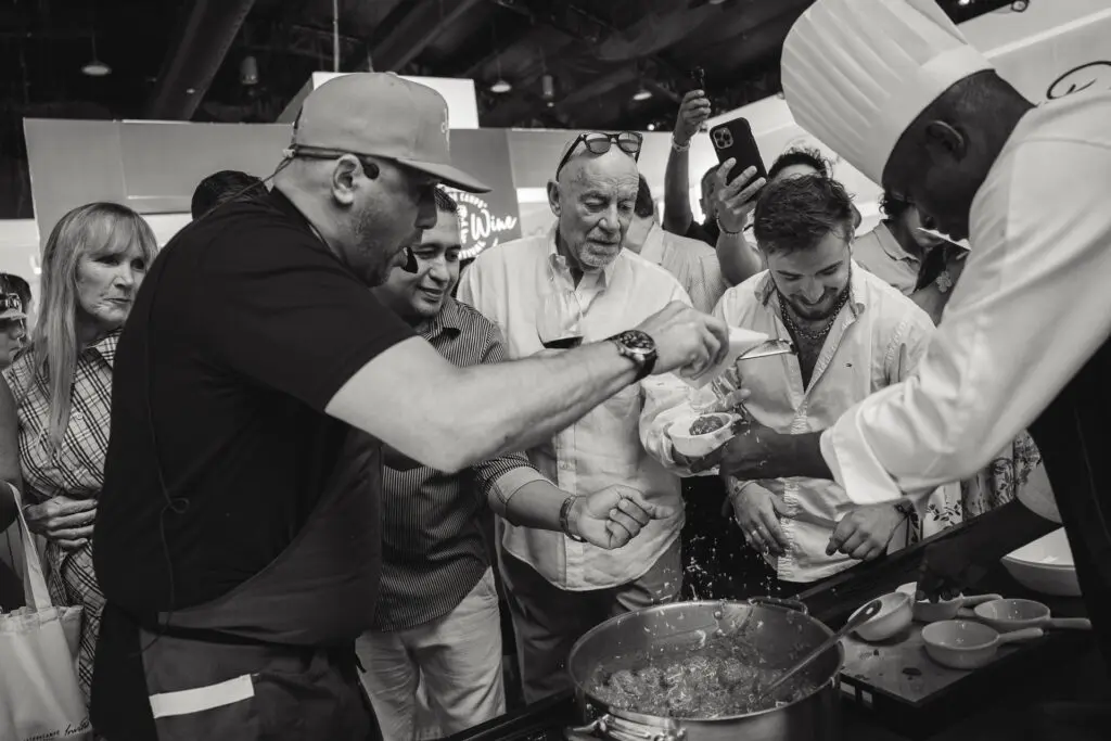 A group of people gathers around a chef cooking, with excitement and engagement as they watch the food preparation.
