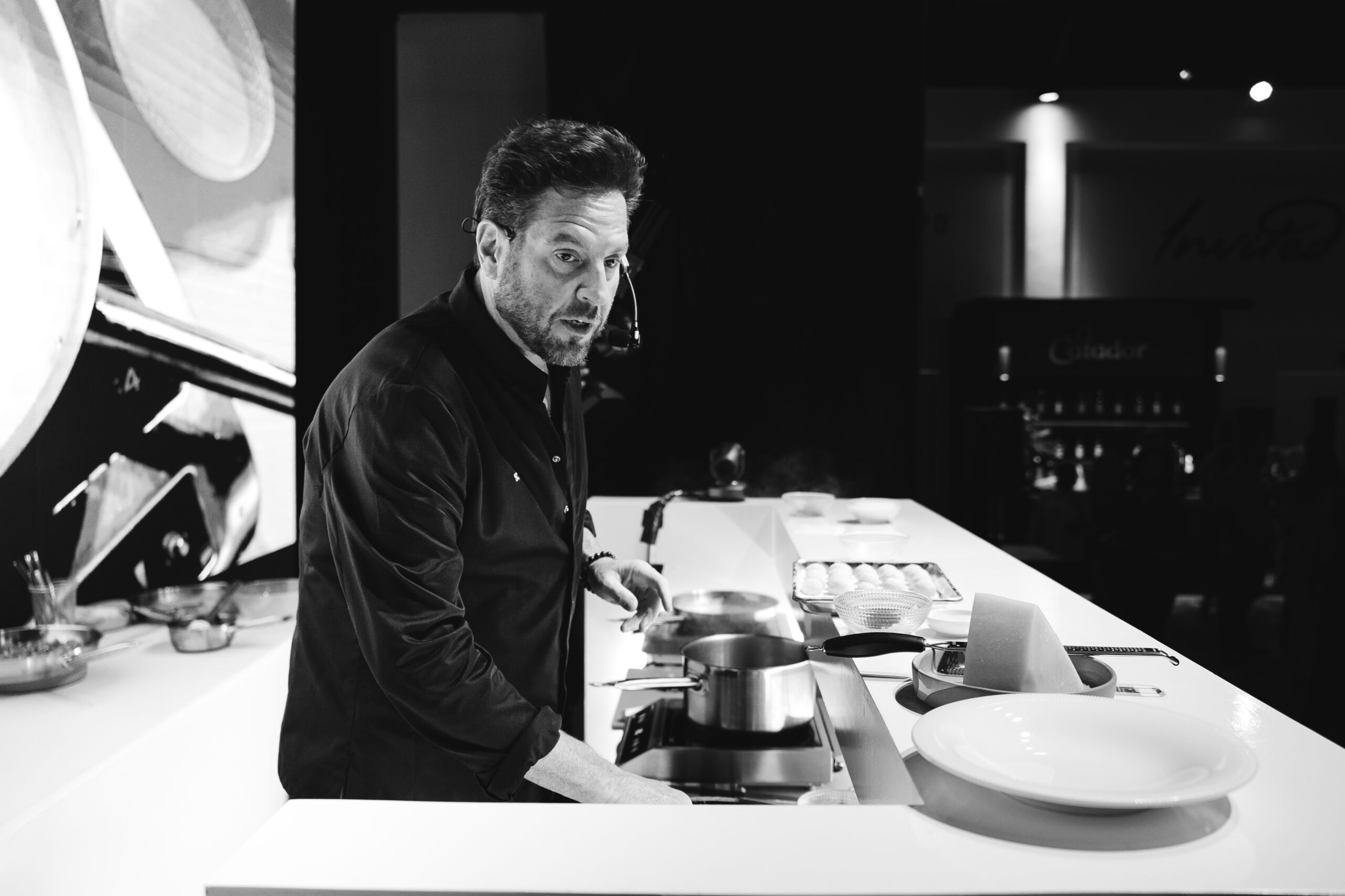  A chef in a black jacket is cooking at a modern kitchen counter, with pots and plates arranged around him. The image is in black and white.