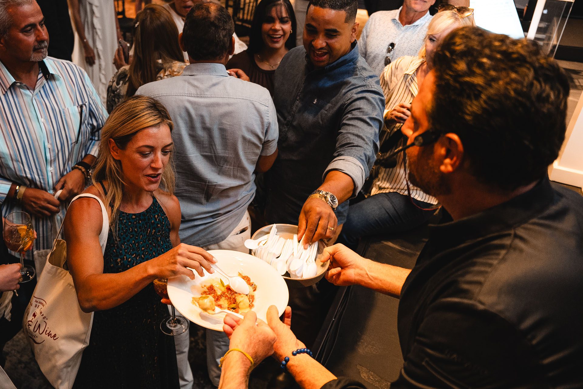  A lively gathering with people enjoying food and drinks, as one woman reaches for a plate while others interact in the background.