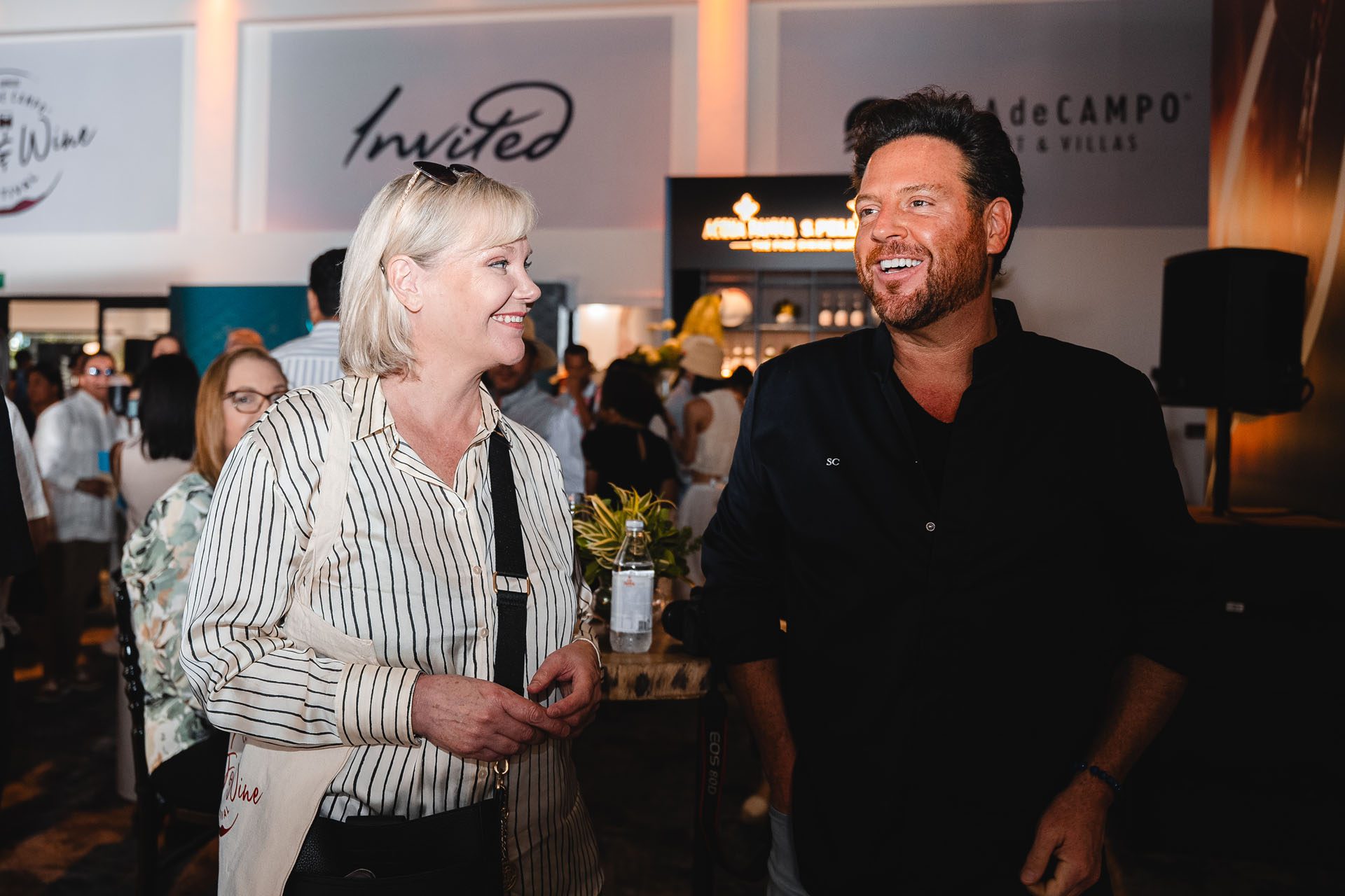 Two people are smiling and engaging in conversation at a lively event, surrounded by a crowd and decorative elements.