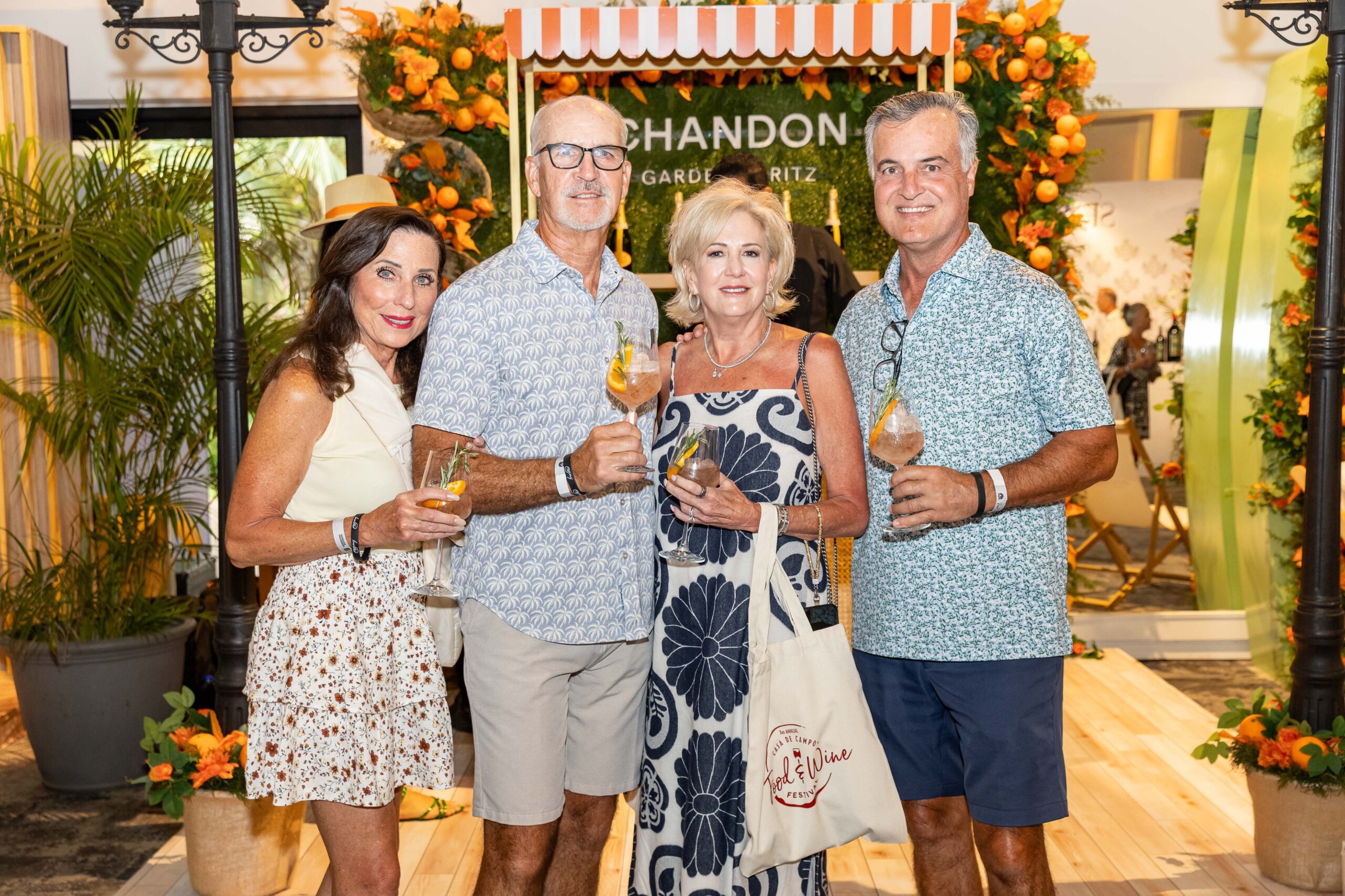 Four people are smiling and holding drinks at a festive event, surrounded by greenery and orange decorations.