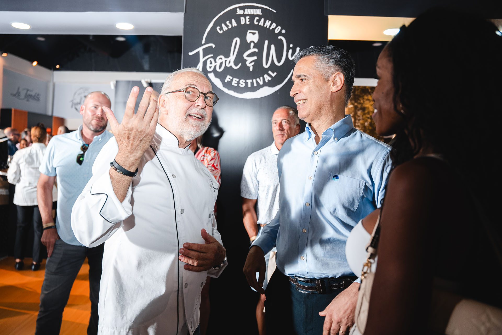  A chef in a white coat gestures while talking to a man in a blue shirt at a food festival event, with others in the background.