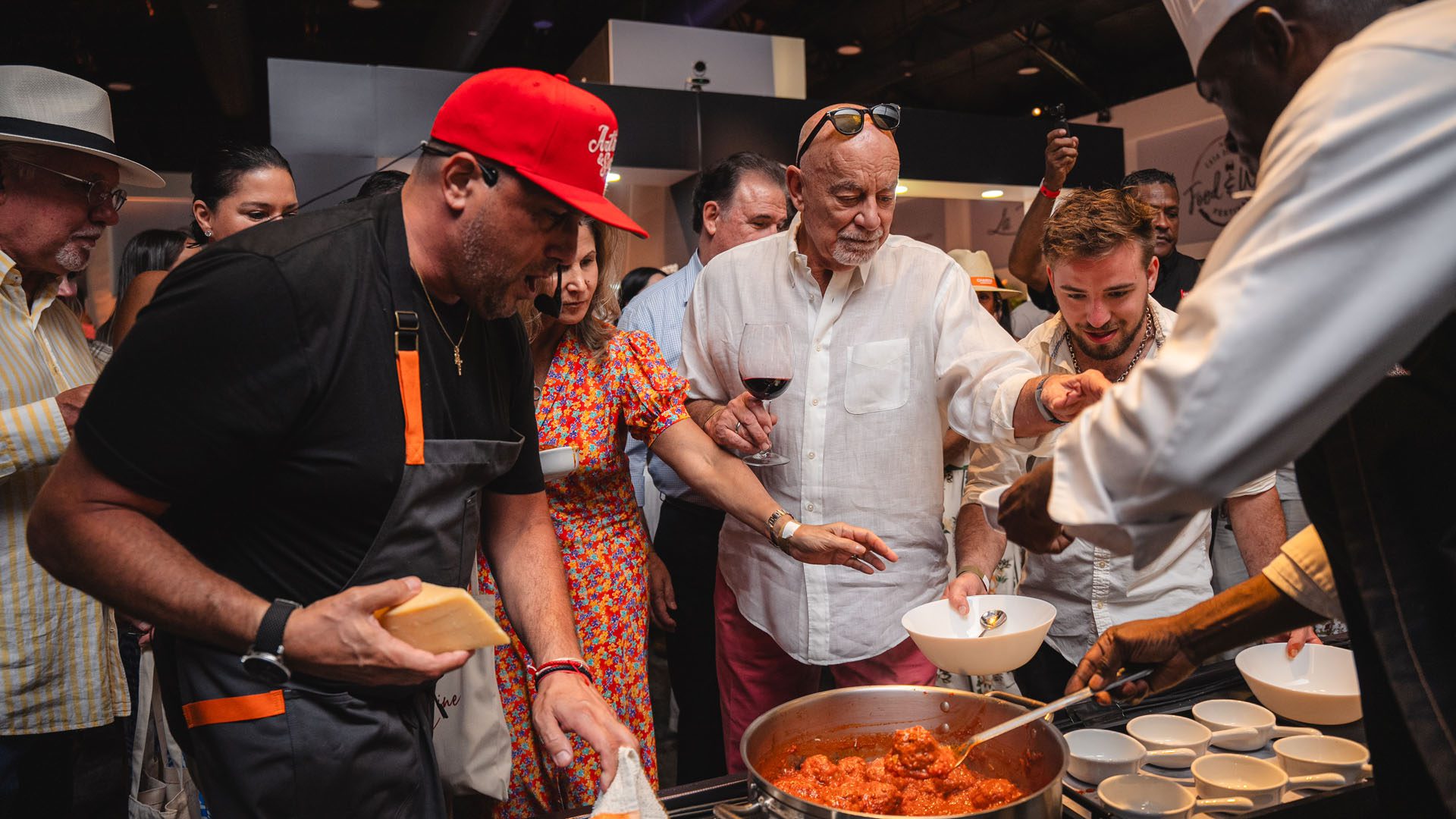 A group of people gathers around a cooking station, enjoying food preparation and serving, with one person holding a glass of wine.