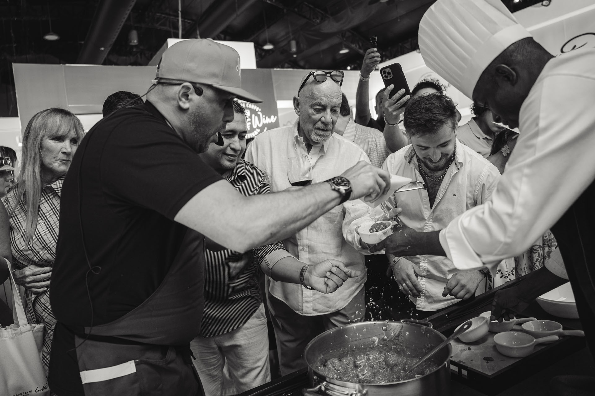 A group of people gathers around a chef cooking, with excitement and engagement as they watch the food preparation.