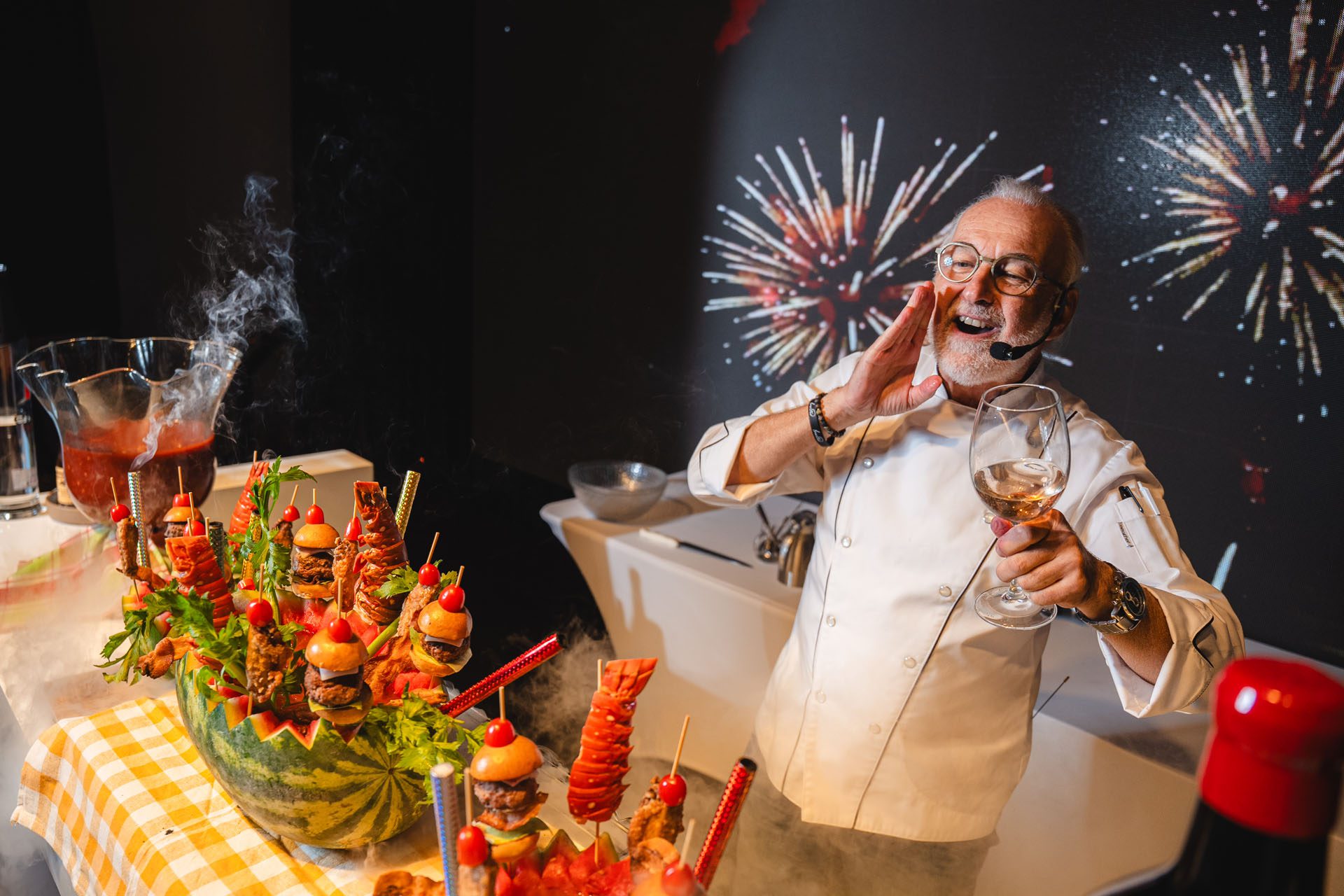 A chef in a white coat joyfully holds a glass while presenting a colorful platter of food, with fireworks displayed in the background.