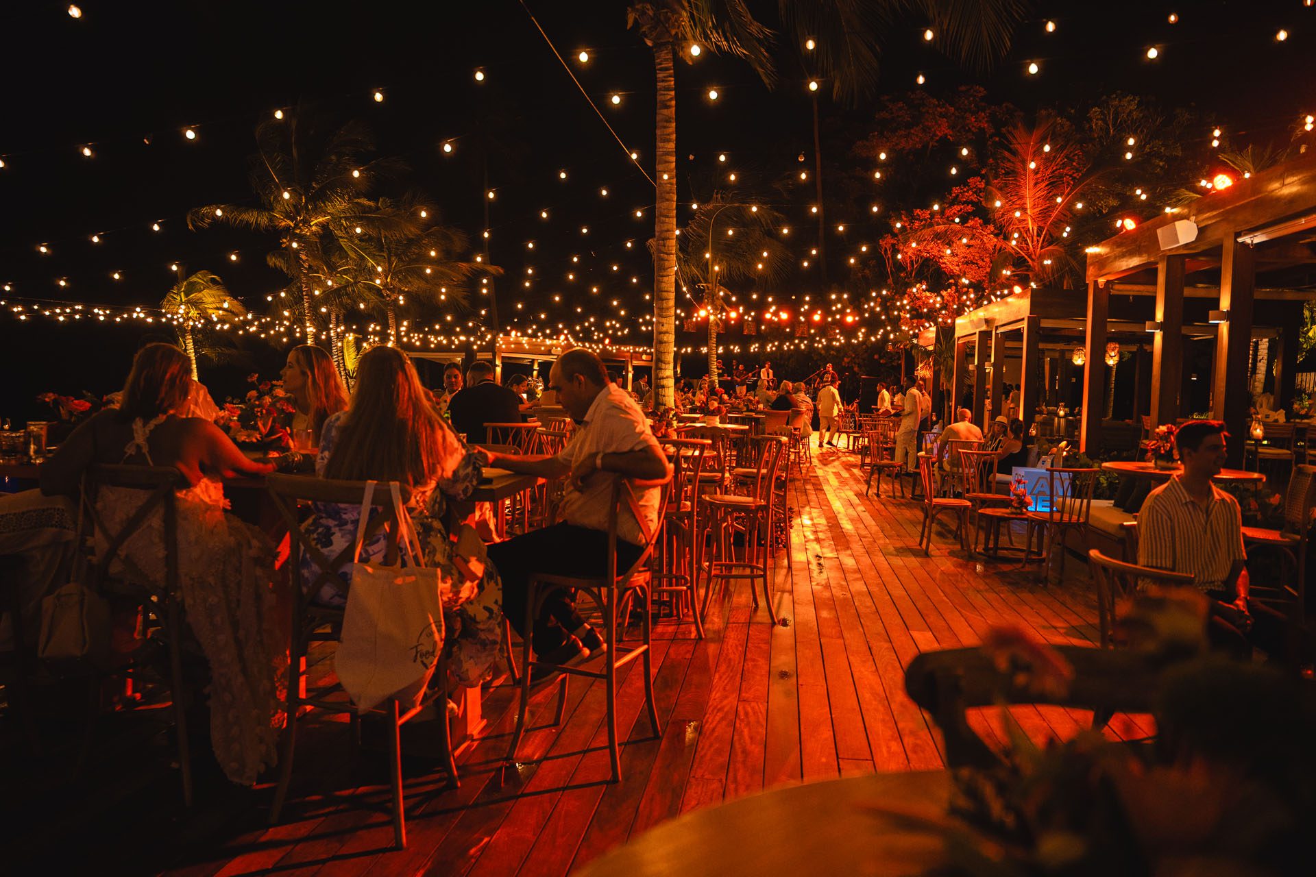 A lively outdoor restaurant at night, adorned with string lights, featuring patrons enjoying drinks and conversation amidst tropical decor.