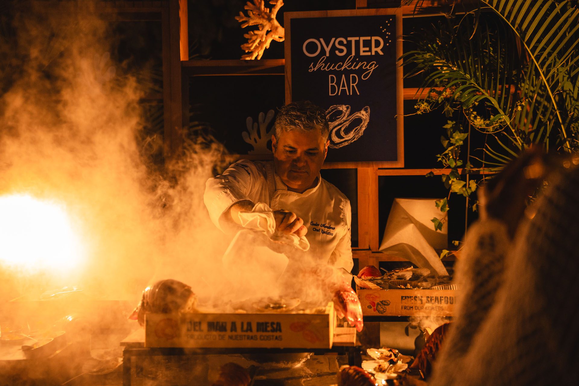  A chef shucks oysters at an oyster bar, surrounded by steam and a tropical setting, with a sign in the background.